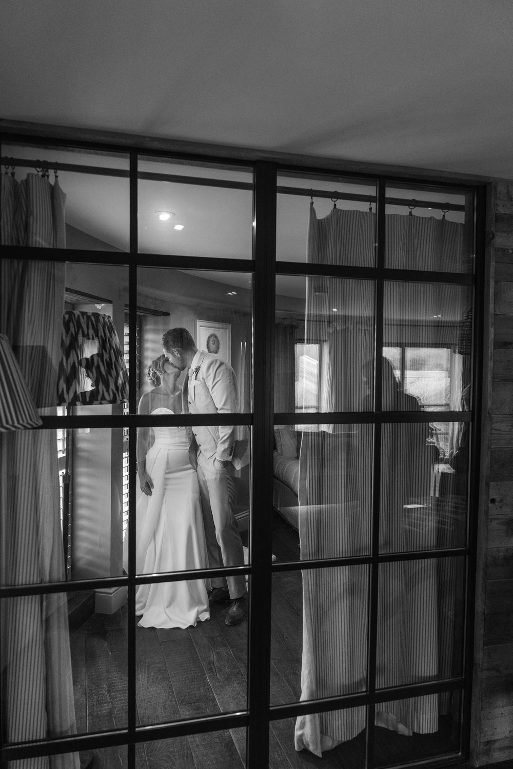 Black and white wedding portrait of a bride and groom sharing a quiet moment behind glass panels in a Gara Rock Hotel suite, photographed du