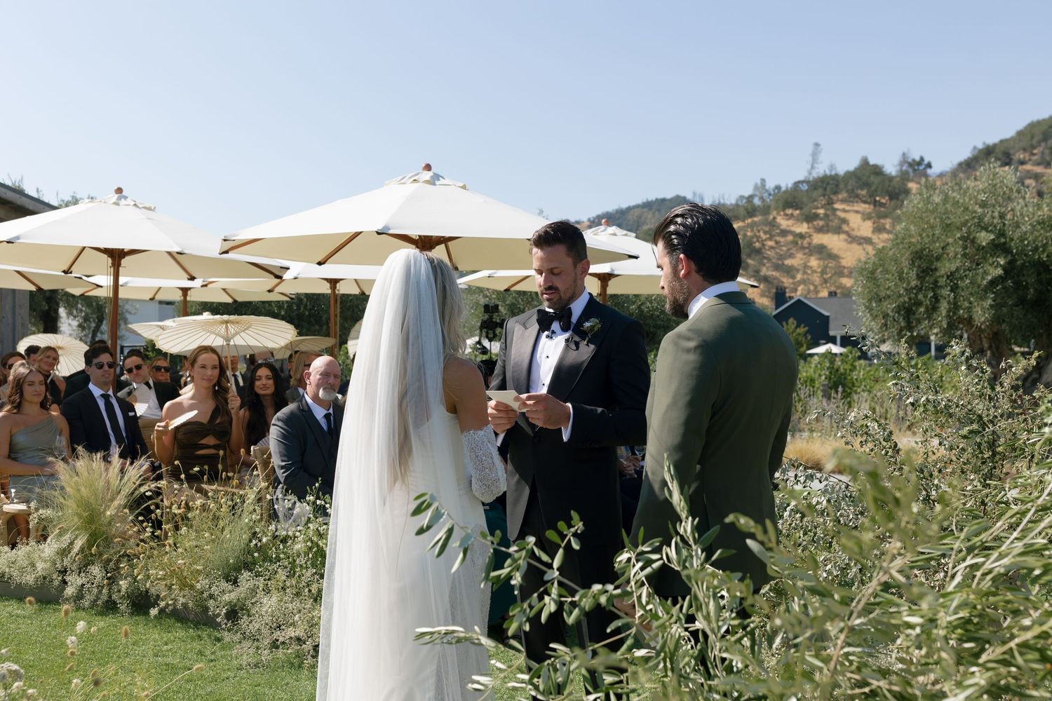 Wedding ceremony guests gather under umbrellas at an outdoor venue on a sunny hillside with white parasols.
