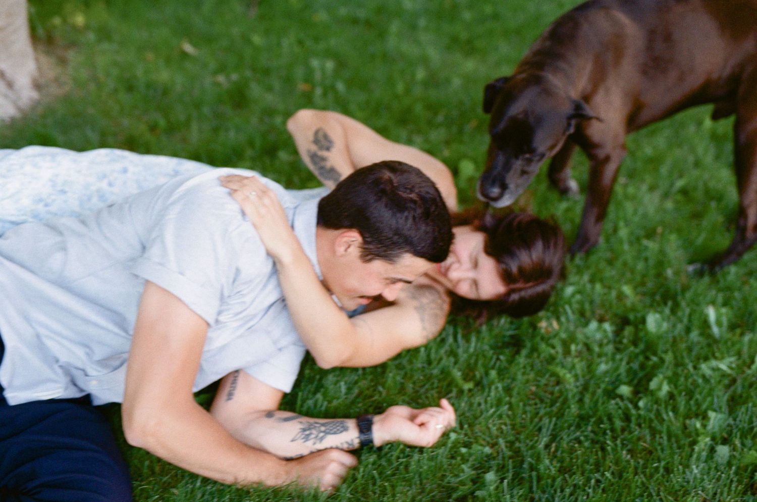 35 mm film photo of couple wrestling playfully in the grass while their dog licks their faces