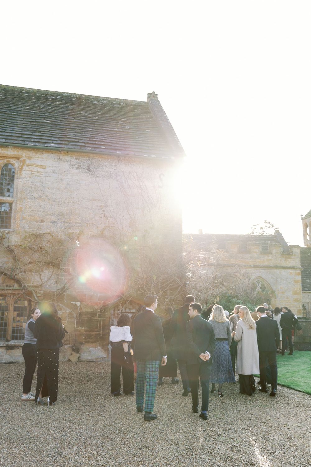 Wedding guests gather outside a rustic stone building on a sunny afternoon during a countryside celebration.
