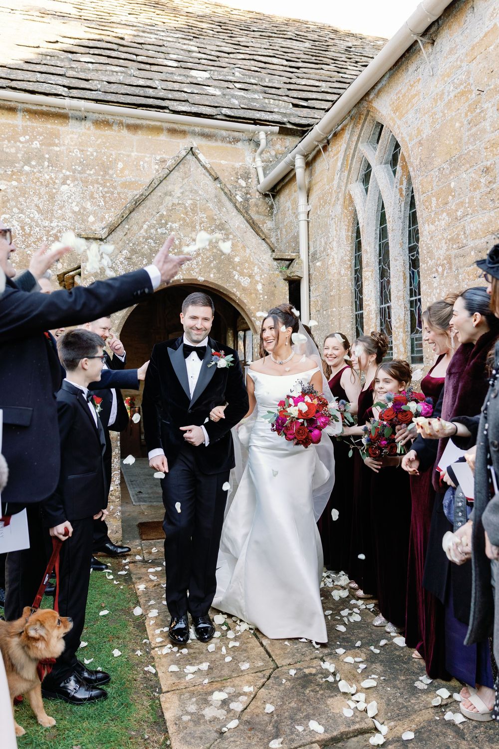 Wedding guests throw white flower petals as newlyweds exit historic stone church building.