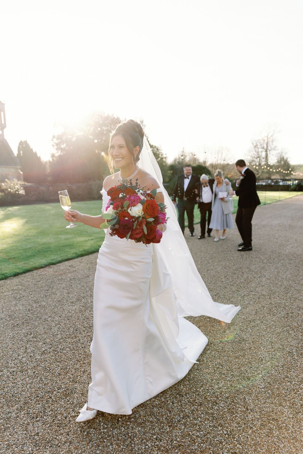 A bride in a white gown holds a red bouquet while walking across a gravel path at sunset.