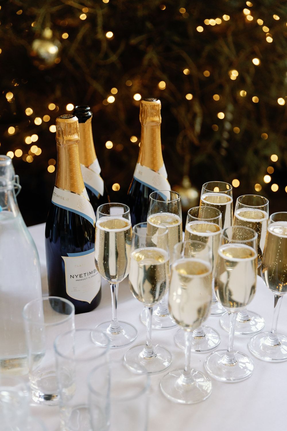 Wedding reception champagne glasses lined up on table with twinkling lights in background.