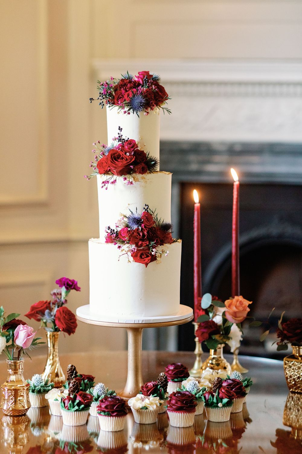 White wedding cake with burgundy floral decorations displayed alongside cupcakes and red candles.