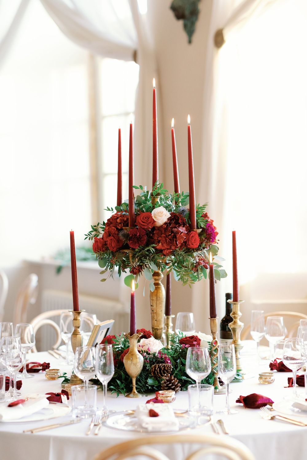 Elegant wedding reception table with red candles, white roses and greenery centerpieces in a bright sunlit room.