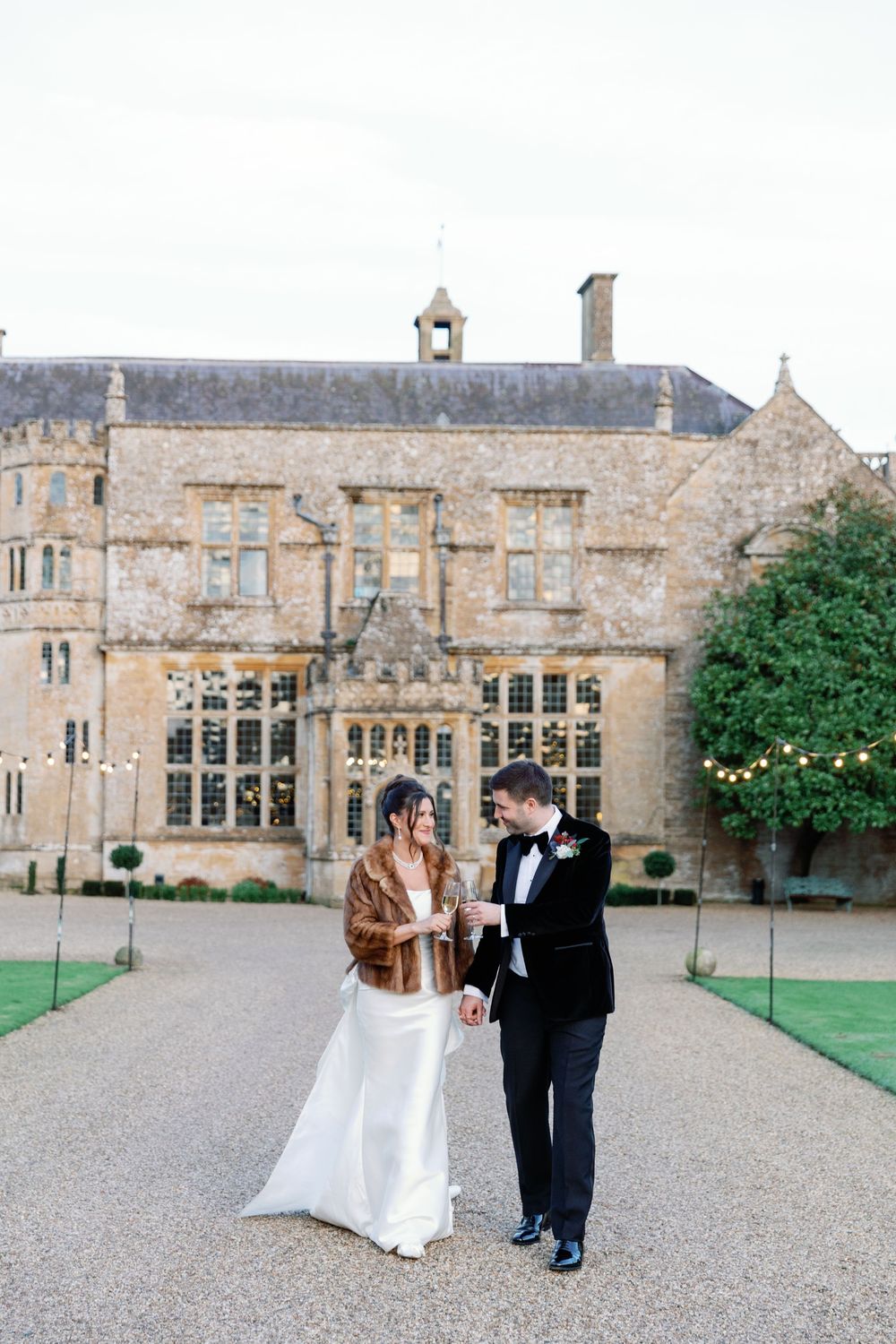 Elegant couple walking outside historic stone mansion during wedding celebration.
