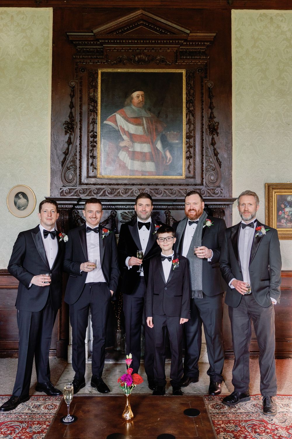 A formal group stands in black tie attire beneath an ornate portrait in a historic room.