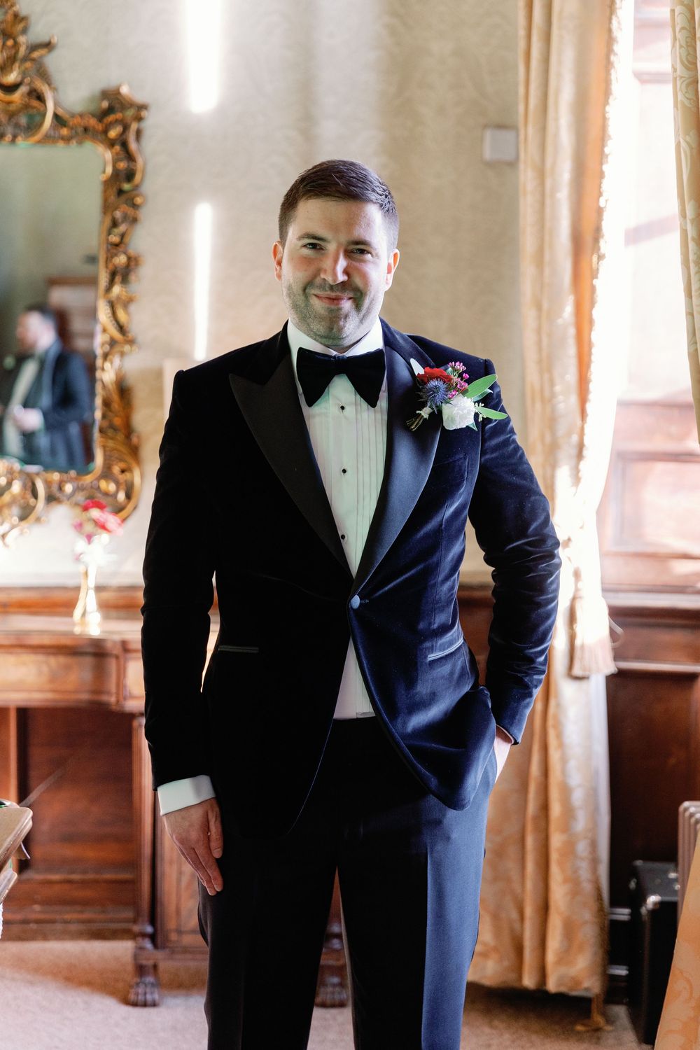 A groom in a classic black tuxedo poses in an elegant room with ornate gold mirrors.