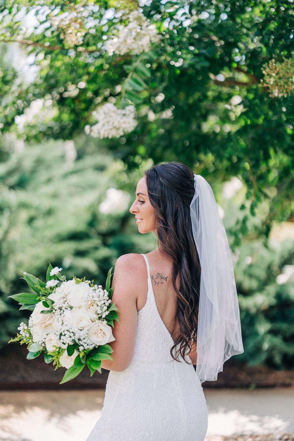 A bride in a white wedding dress holds a bouquet of white flowers while posing in a garden setting at The Barn South