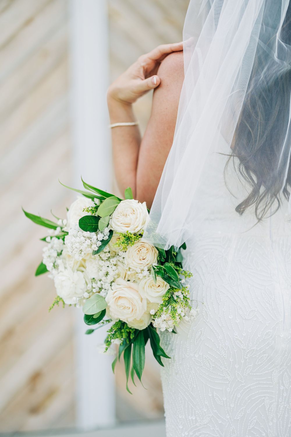 Close-up details of white lace wedding dress fabric with delicate floral bouquet featuring white roses and greenery at The Barn South