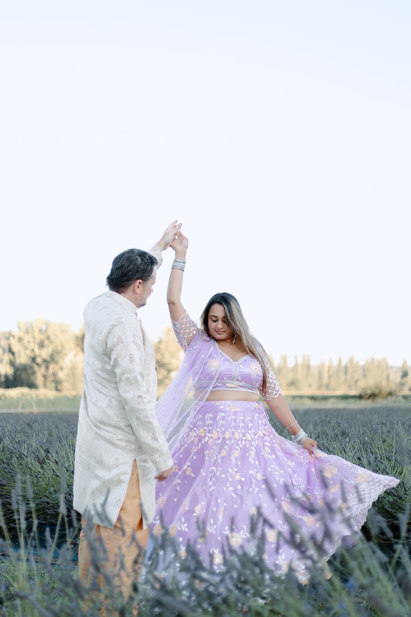 A couple dances in a field at sunset, with the woman wearing a flowing lavender lehenga dress that twirls gracefully.