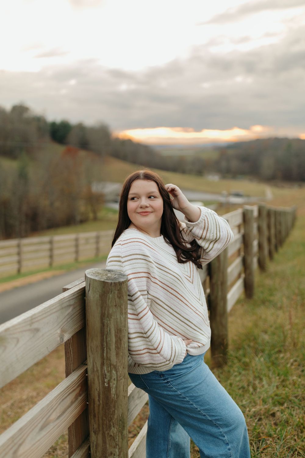 Person in a white striped sweater and jeans poses casually against a wooden fence in a rural countryside setting at dusk.