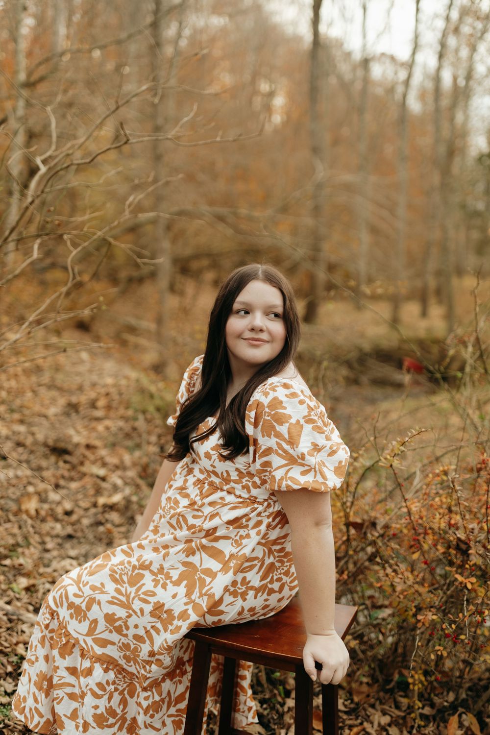 A person in a floral dress sits on a wooden stool in an autumn forest setting with fallen leaves and bare trees.
