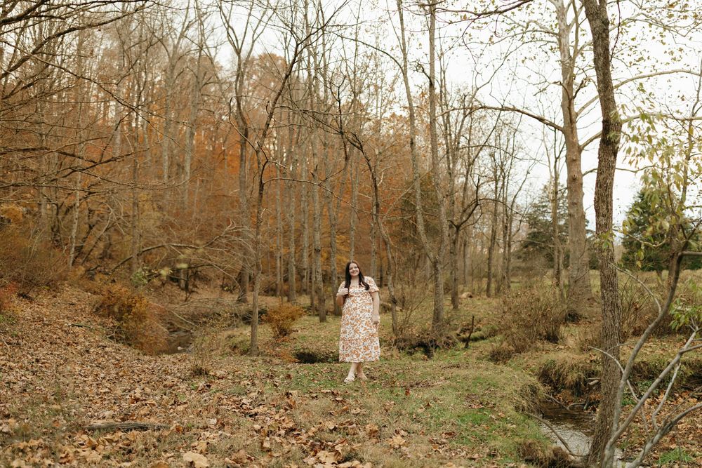 A figure in a white dress stands on a leaf-covered path in an autumn forest clearing.
