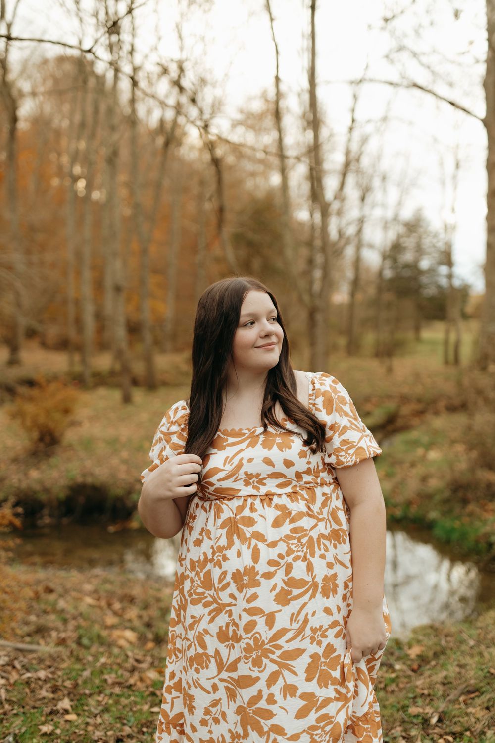 A floral dress in orange and white tones complements the autumn scenery with bare trees and a stream in the background.