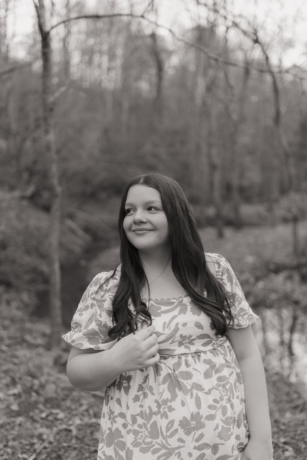A person wearing a floral dress poses playfully in a black and white photo series outdoors among trees and fallen leaves.