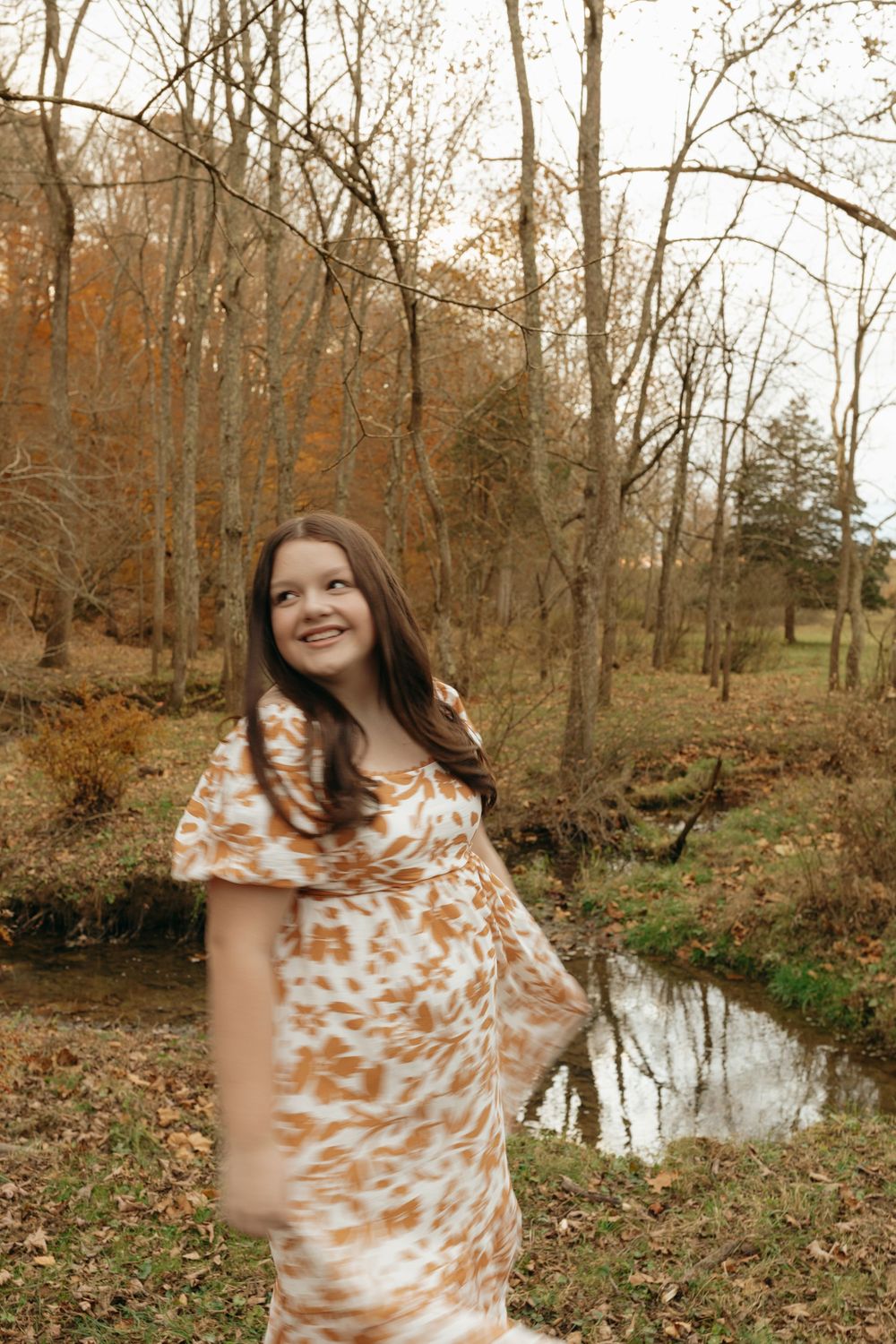 A joyful figure spins in a flowing dress against a backdrop of bare trees and a peaceful stream.