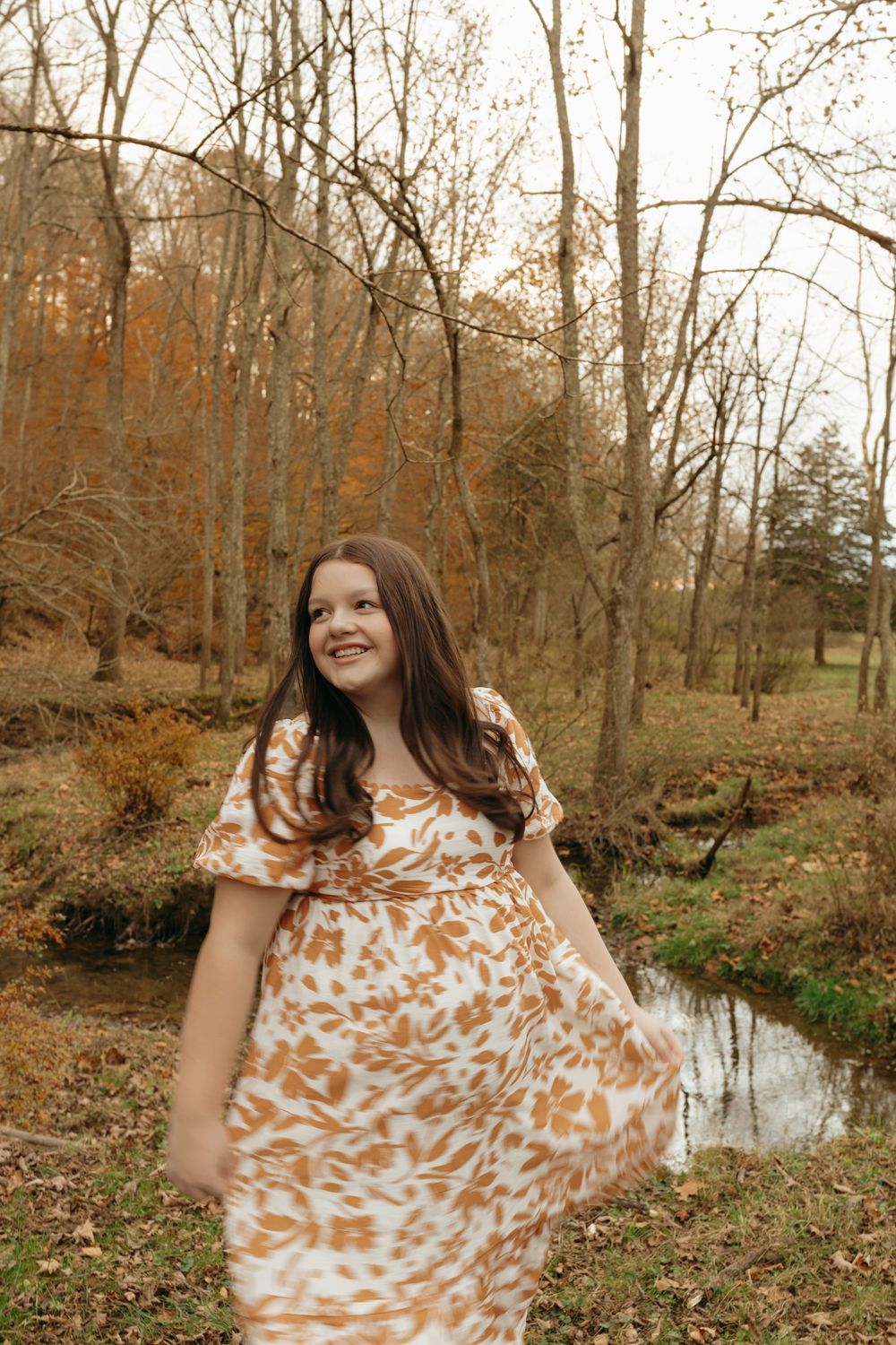 A person in a floral dress twirls playfully by a stream in an autumn forest setting.