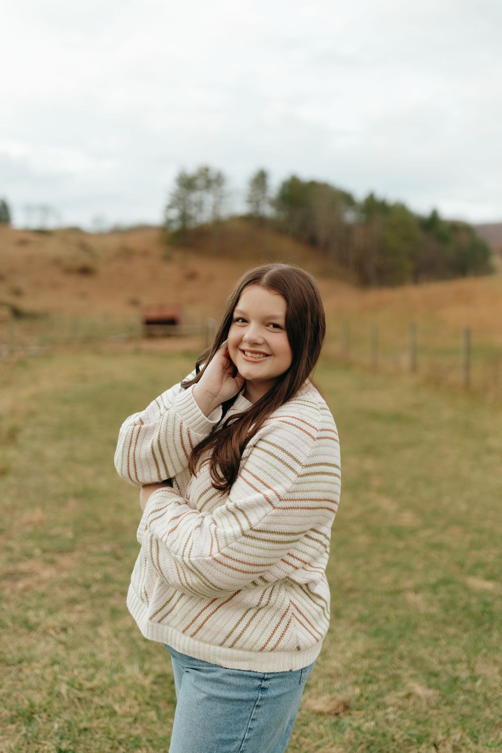 A casual outdoor portrait featuring a striped sweater and jeans against a rural hillside backdrop.