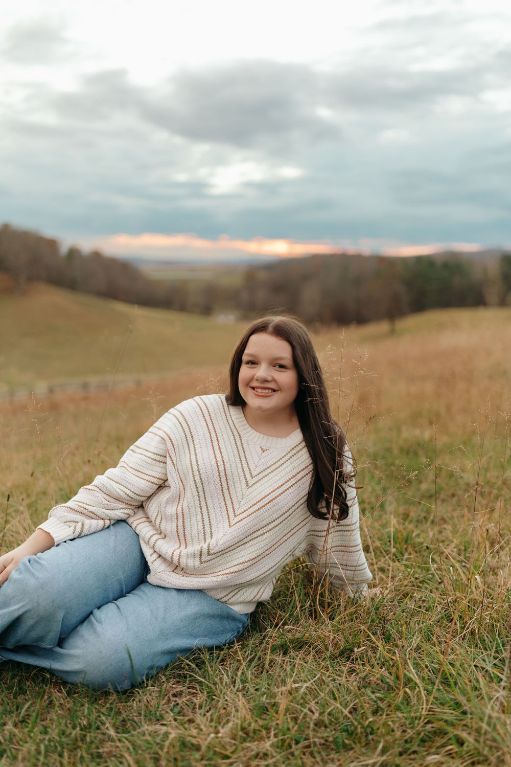 A person in a striped sweater and jeans relaxes in a golden autumn field during an outdoor photo session.