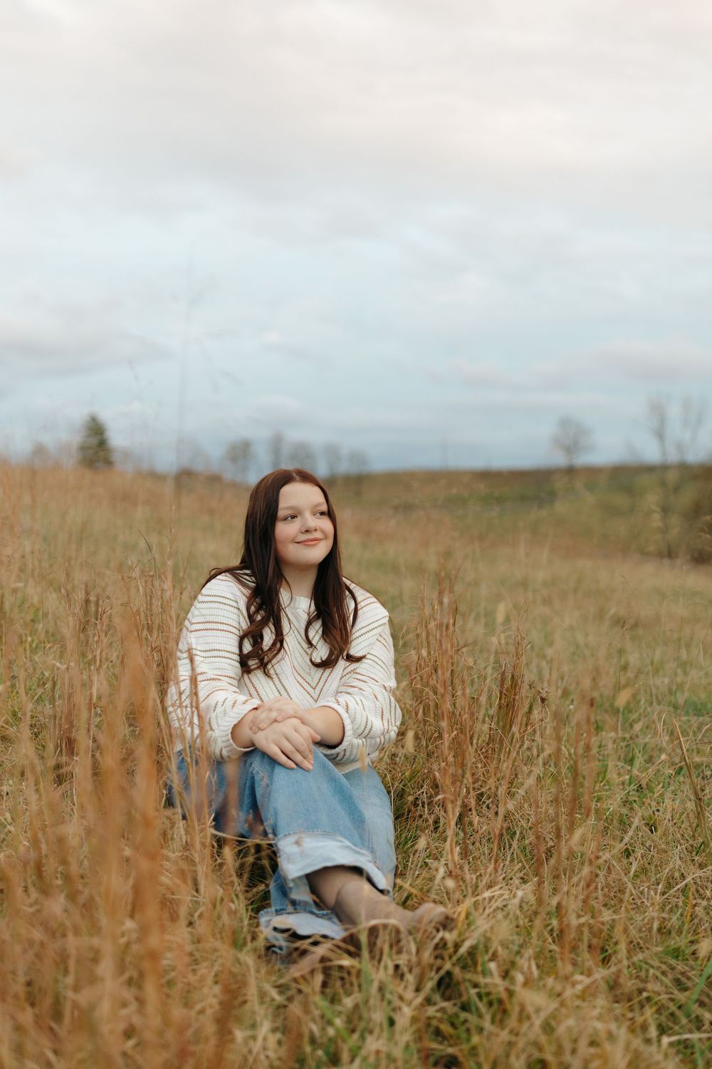 Someone sits among tall dried grass in a field on an overcast autumn day wearing denim and white.