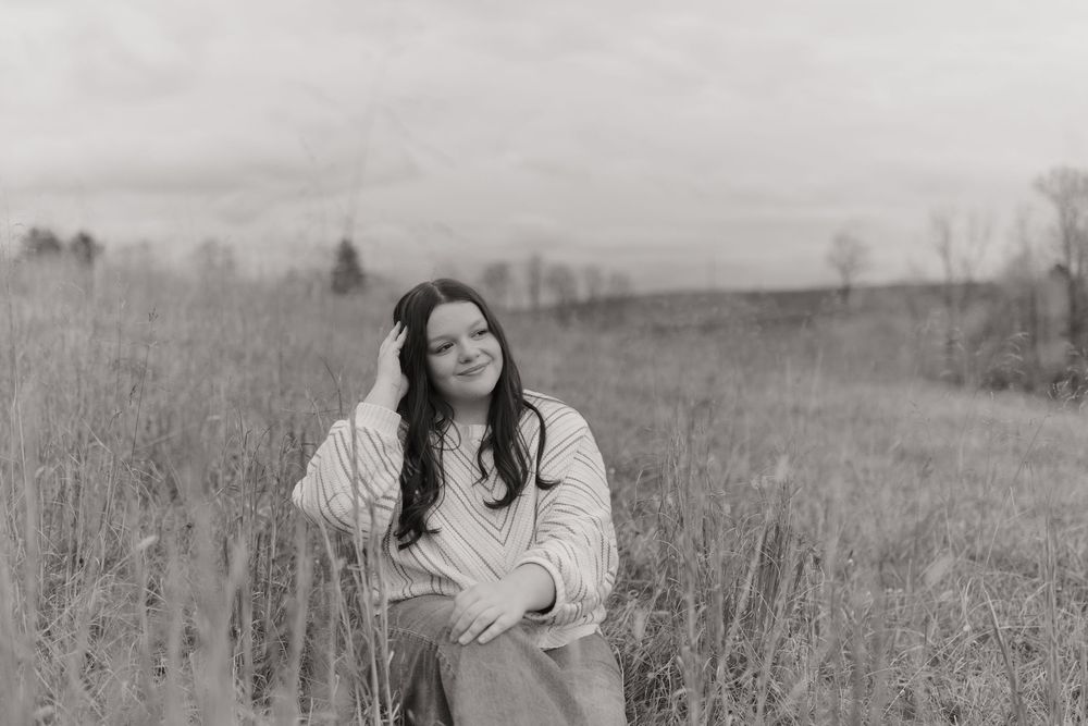 A woman in a white dress sits in a field of tall grass during a black and white outdoor photo session.