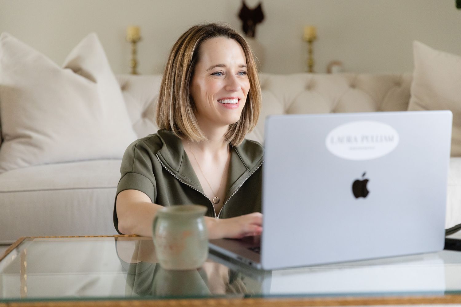 Photographer works on a MacBook laptop while sitting on a couch with throw pillows in a bright, cozy home office setting in Spring Texas