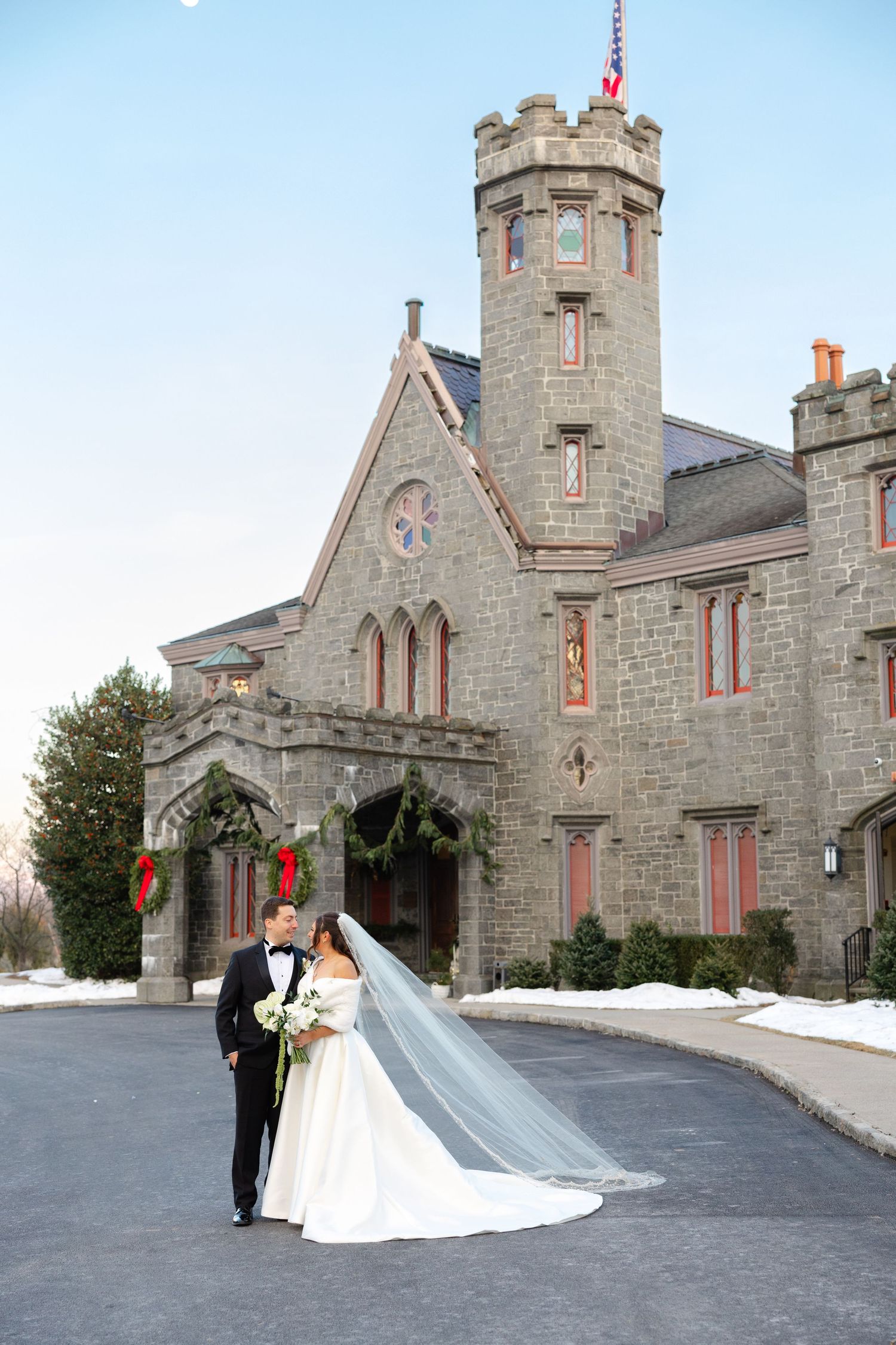 A couple stands outside in front of the Whitby Castle in Rye, NY in wedding attire on their winter wedding day.