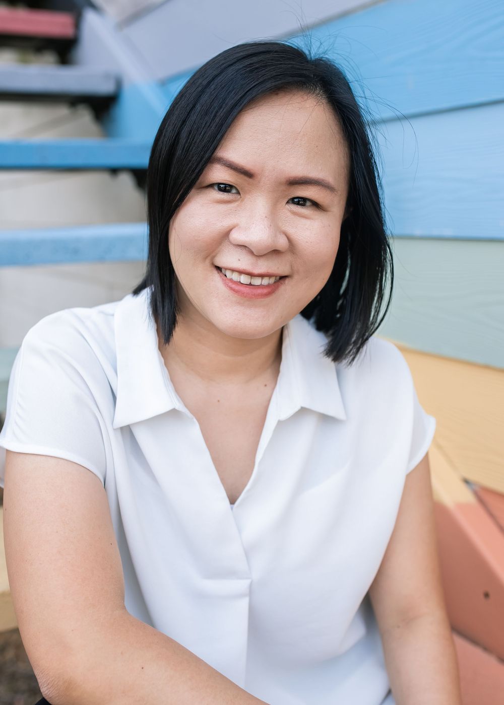A person in a white collared shirt smiles warmly while sitting against a colorful wall background.