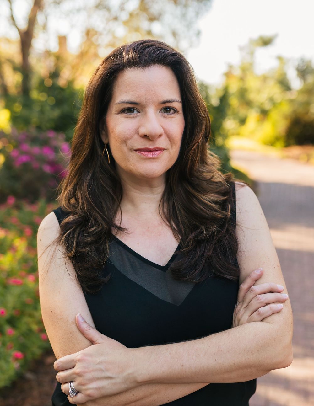 A person in a black sleeveless top poses outdoors with greenery and flowers in the background during golden hour.