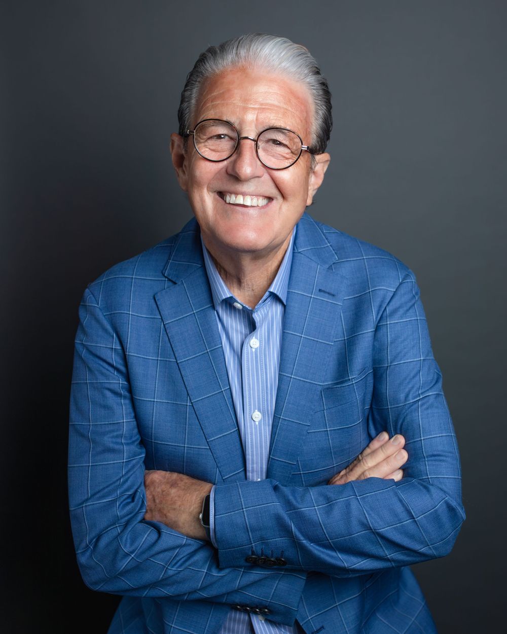Professional headshot portrait of someone wearing a blue checkered blazer and glasses against a dark background.