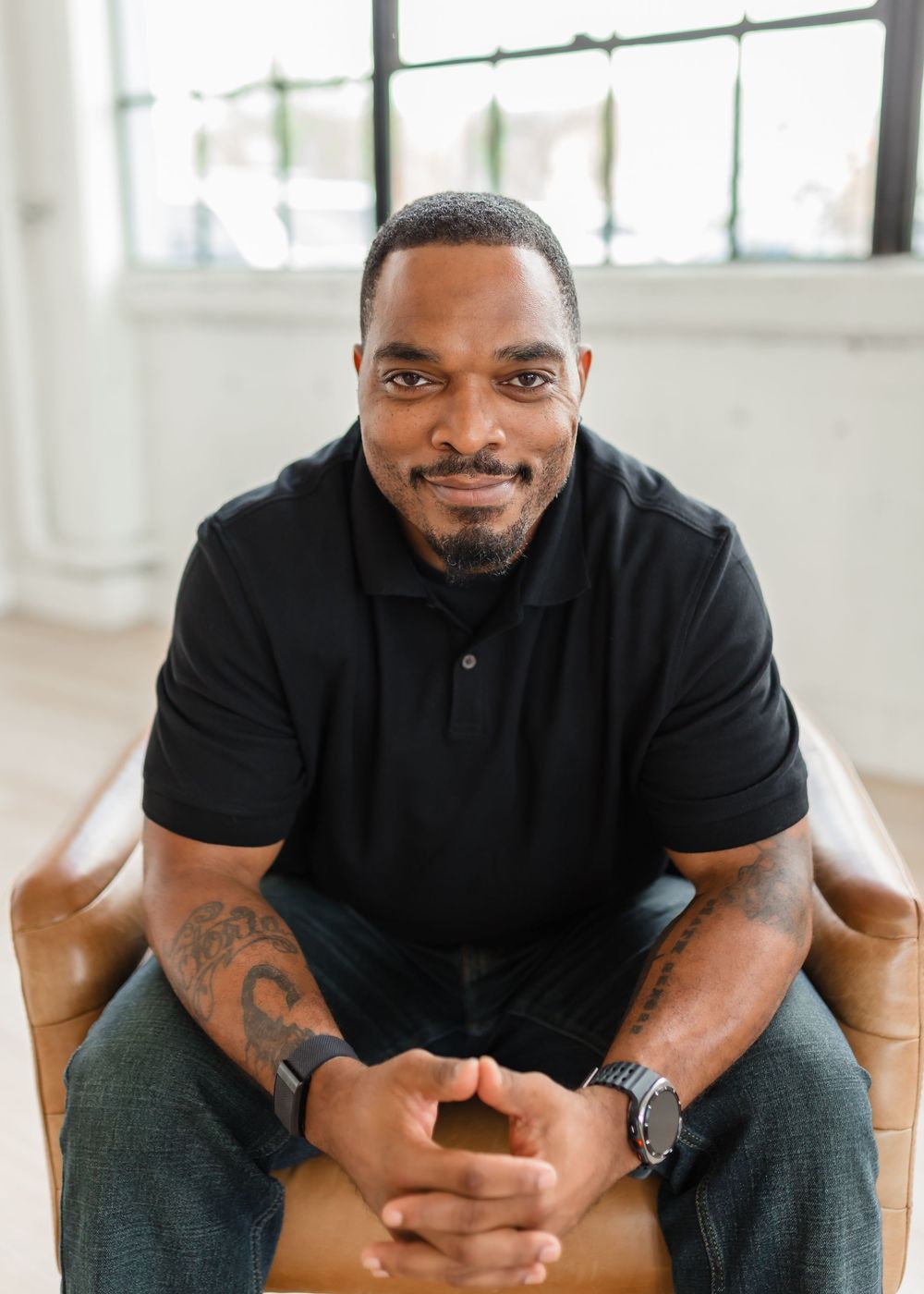 A person in a black polo shirt sits casually in a tan chair, with clasped hands and visible arm tattoos for headshots.