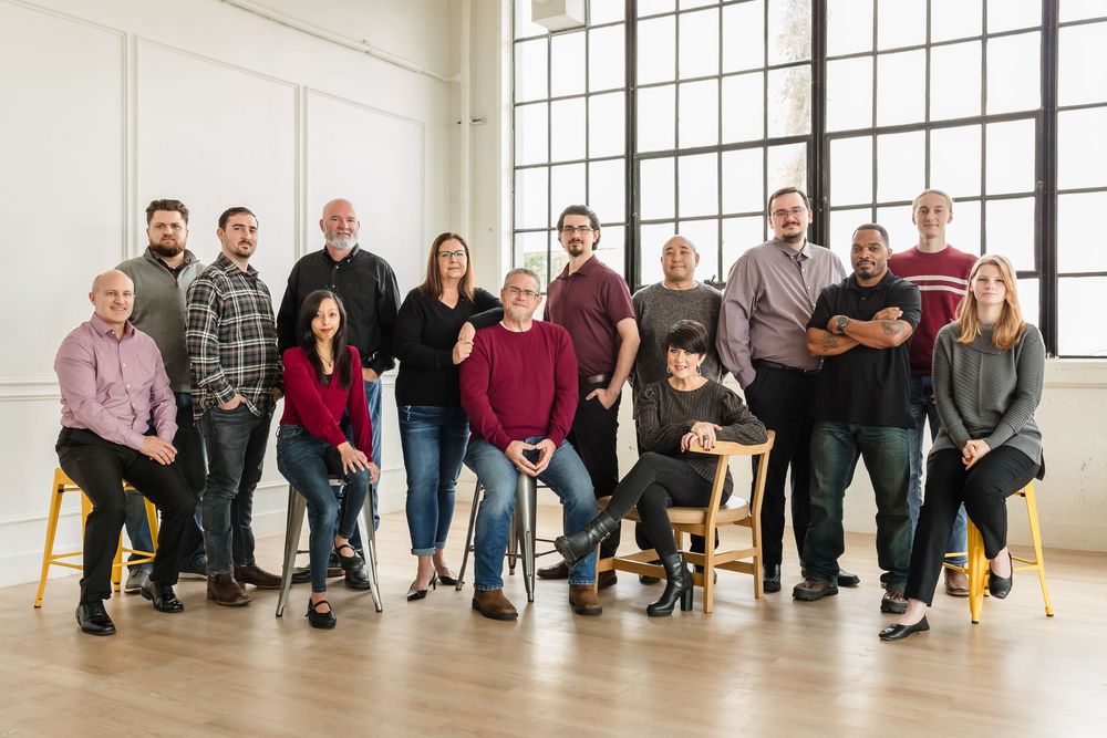 A panoramic group photo of people sitting casually in a bright, sunlit studio with large windows for Business Headshots.
