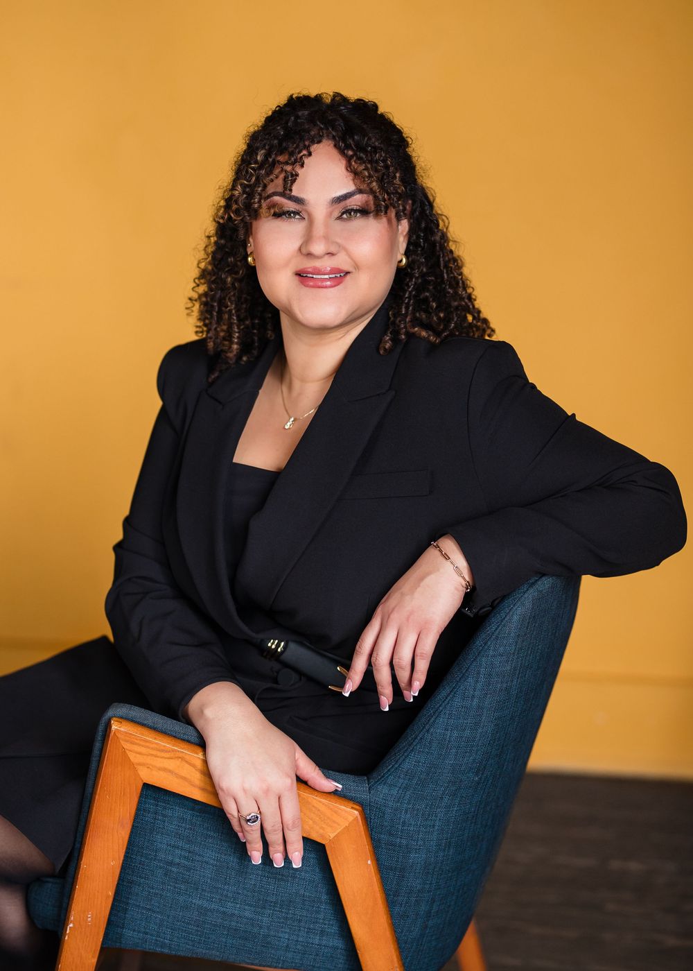 Professional portrait of someone in a black blazer and jeans seated on a wooden chair against a golden yellow backdrop for Headshots.