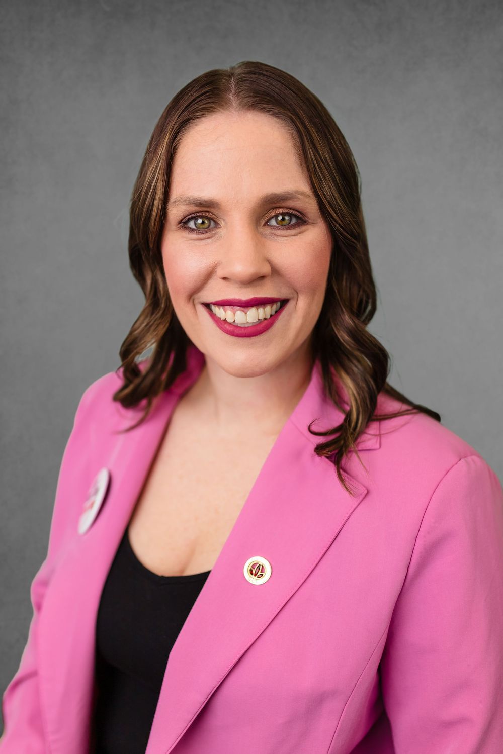 Professional headshot portrait of someone in a bright pink blazer and black top against a gray background.