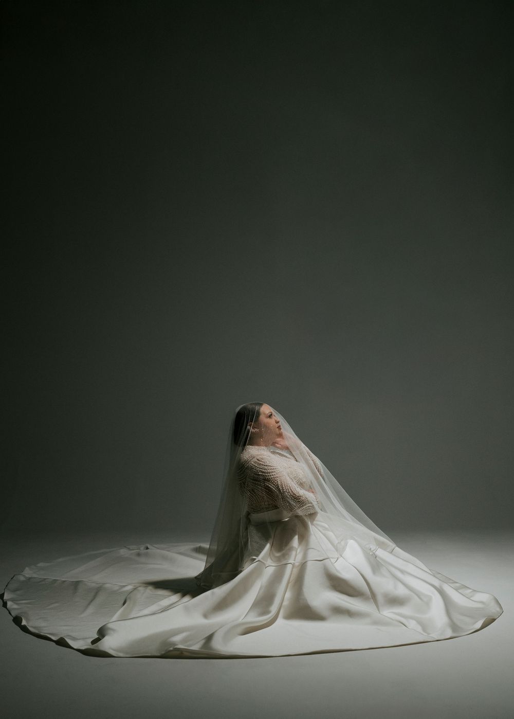 A figure in a flowing white wedding dress lies gracefully on a dark studio floor creating an artistic bridal portrait.