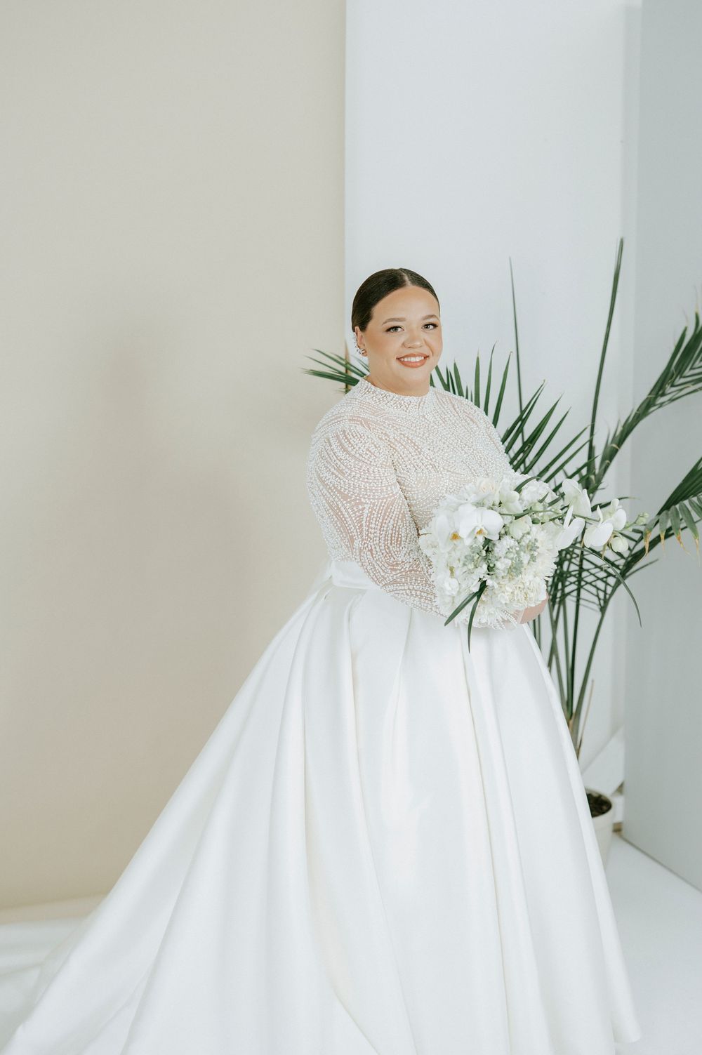 A bride in a long-sleeved lace and satin ball gown holds a white floral bouquet next to tropical palm leaves.