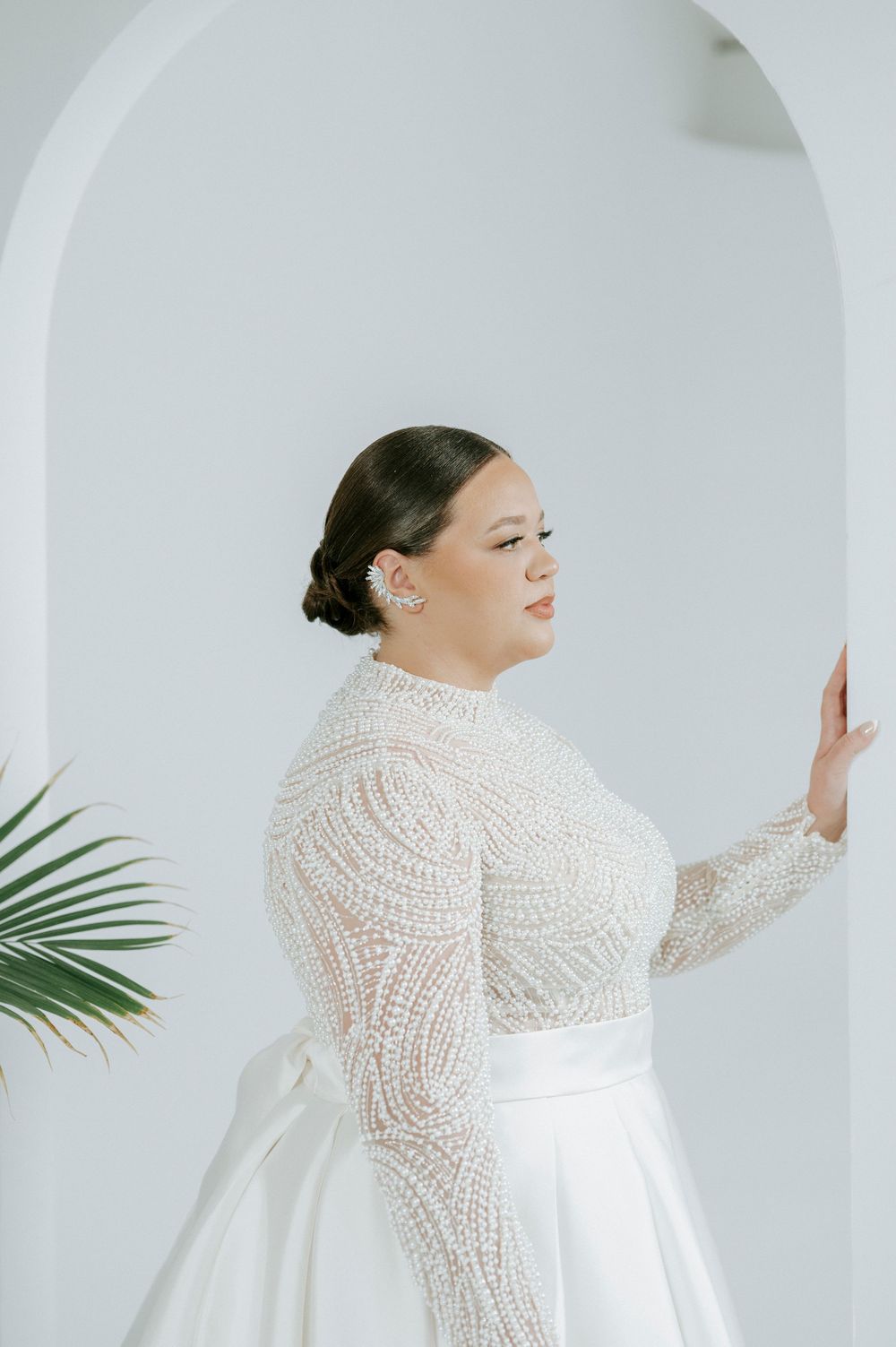 A bride in a long-sleeved lace wedding dress with a high neckline poses against a white backdrop with palm fronds.