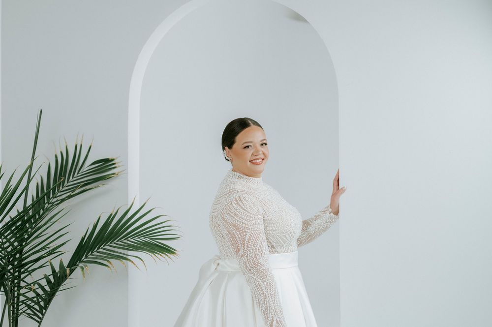 A figure in a flowing white lace wedding gown poses gracefully against a light gray wall with palm fronds.
