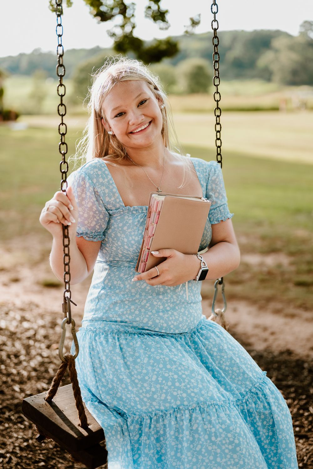 Someone in a blue floral dress sits peacefully on a swing while reading from a tablet device.