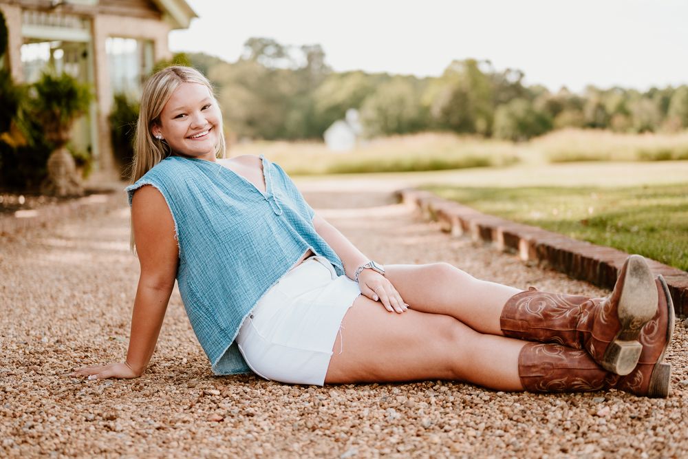 A person in a sleeveless blue top and white shorts relaxes on a gravel path wearing brown boots in a rural setting.