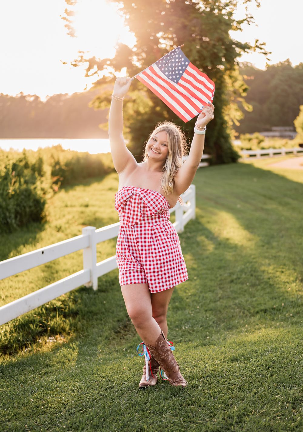 Patriotic summer scene with American flag being waved at sunset by a white fence with cowboy boots and gingham dress.