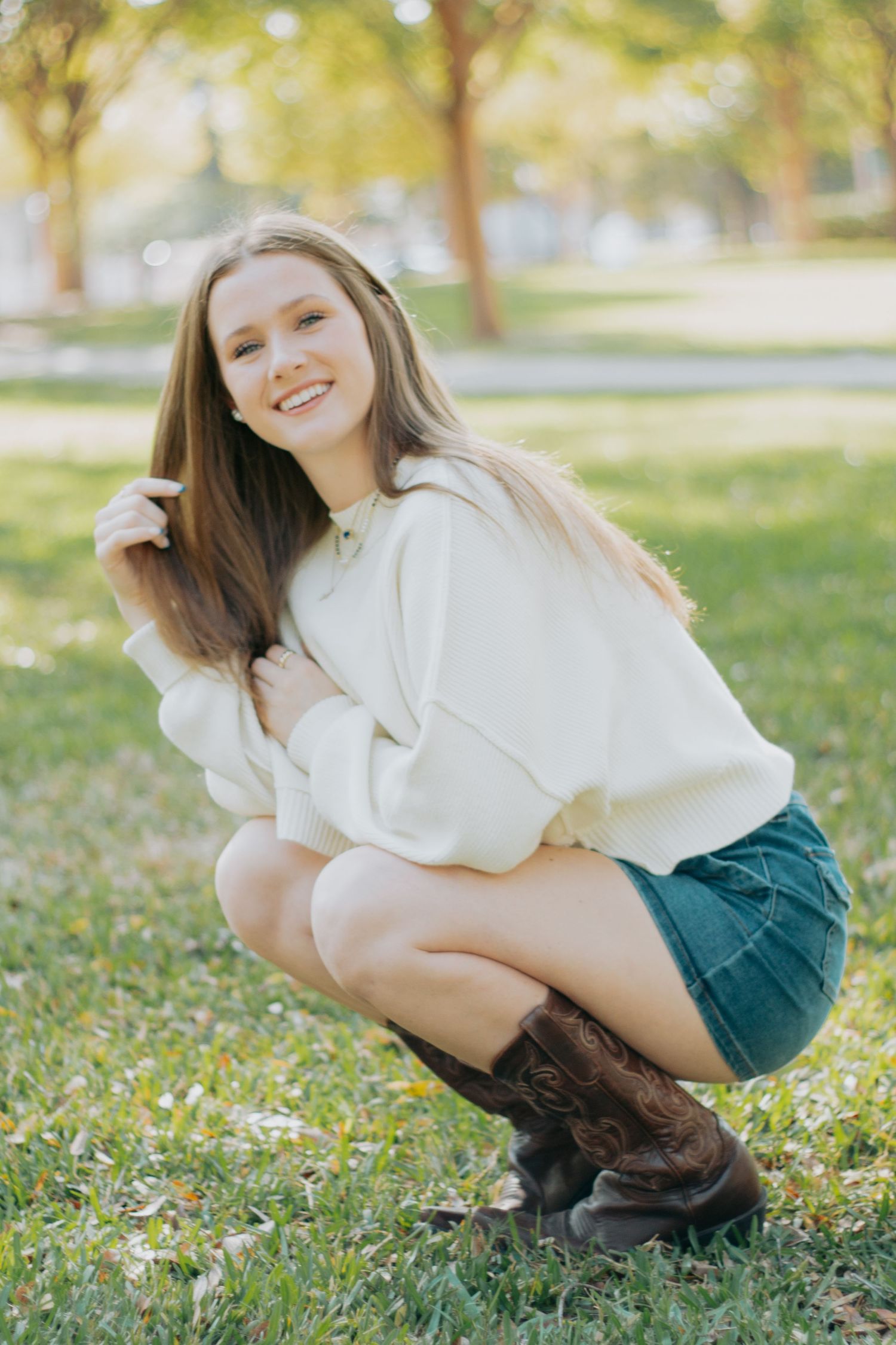 A person in a white sweater and green skirt crouches playfully in the grass on a sunny day.