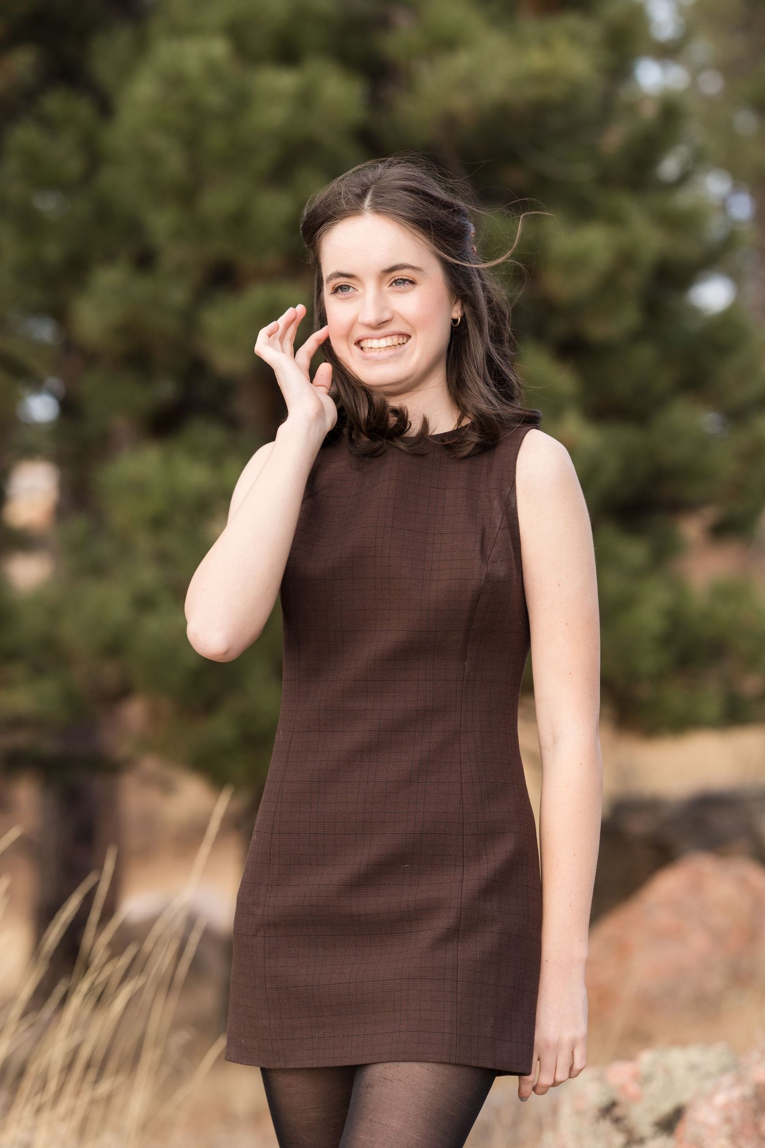 Professional headshot portrait of someone in a sleeveless brown dress against a blurred outdoor background.