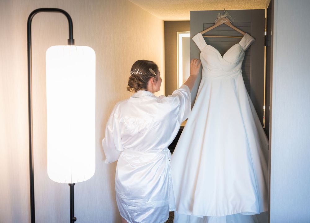 Bride in white robe admiring her wedding dress hanging on a door next to a modern floor lamp.