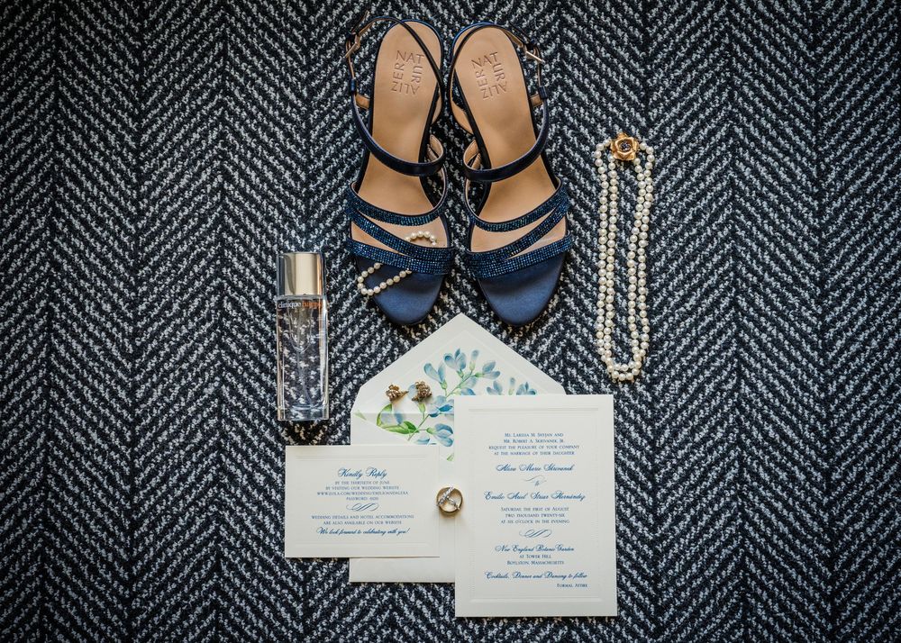 Navy blue wedding heels, perfume, pearl necklace, and floral wedding invitation flat lay display.