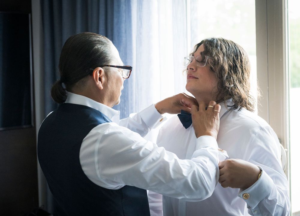 Groom getting bow tie adjusted by man near window during wedding day preparations indoors.