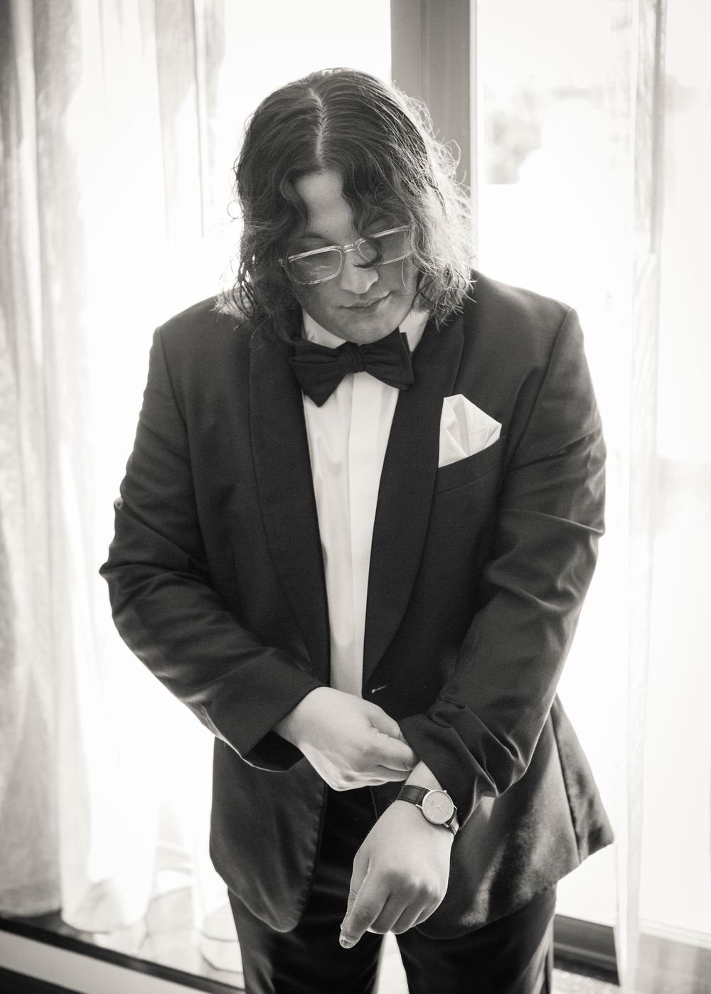 Black and white photo of a man in a tuxedo with bow tie and glasses, adjusting his watch near a window.