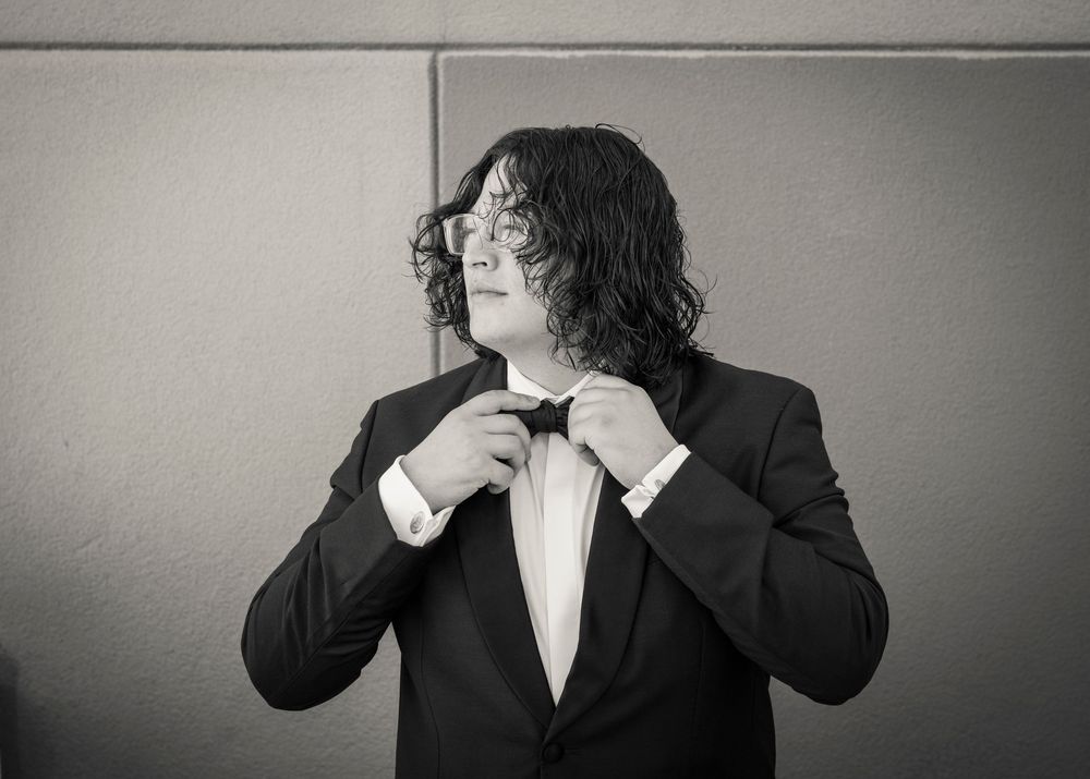 Man in black tuxedo adjusting bow tie, long curly hair and glasses, black and white formal portrait photo.