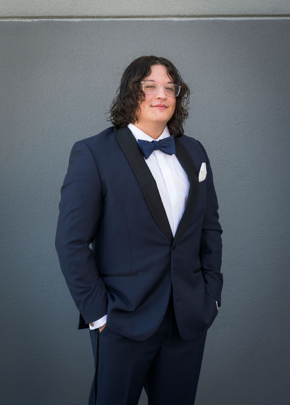Groom in navy tuxedo with bow tie and white pocket square posing against gray wall.